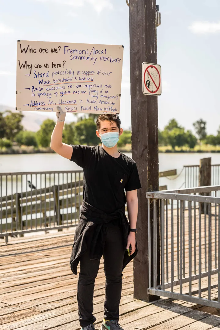 A masked person on a waterfront pier holding a hand-lettered sign that begins: Who are we? Fremont and local community members.