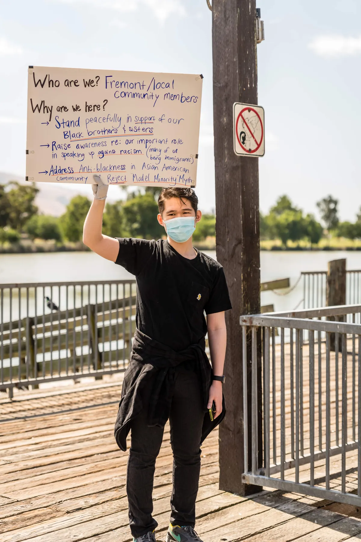 A masked person on a waterfront pier holding a hand-lettered sign that begins: Who are we? Fremont and local community members.