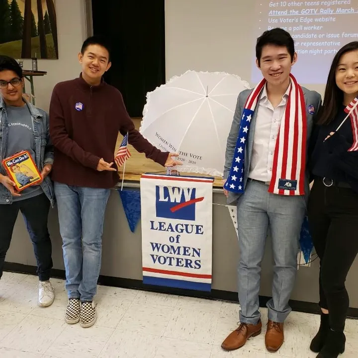 Four students beside a League of Women Voters banner and a parasol reading Women Win the Vote, 1920 to 2020.