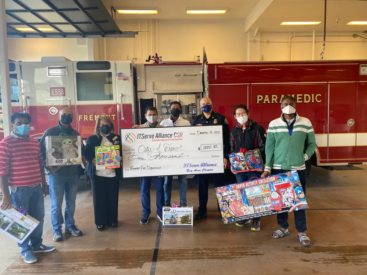 People in front of a Fremont fire engine holding a presentation cheque and boxed toys, beside a firefighter in uniform.