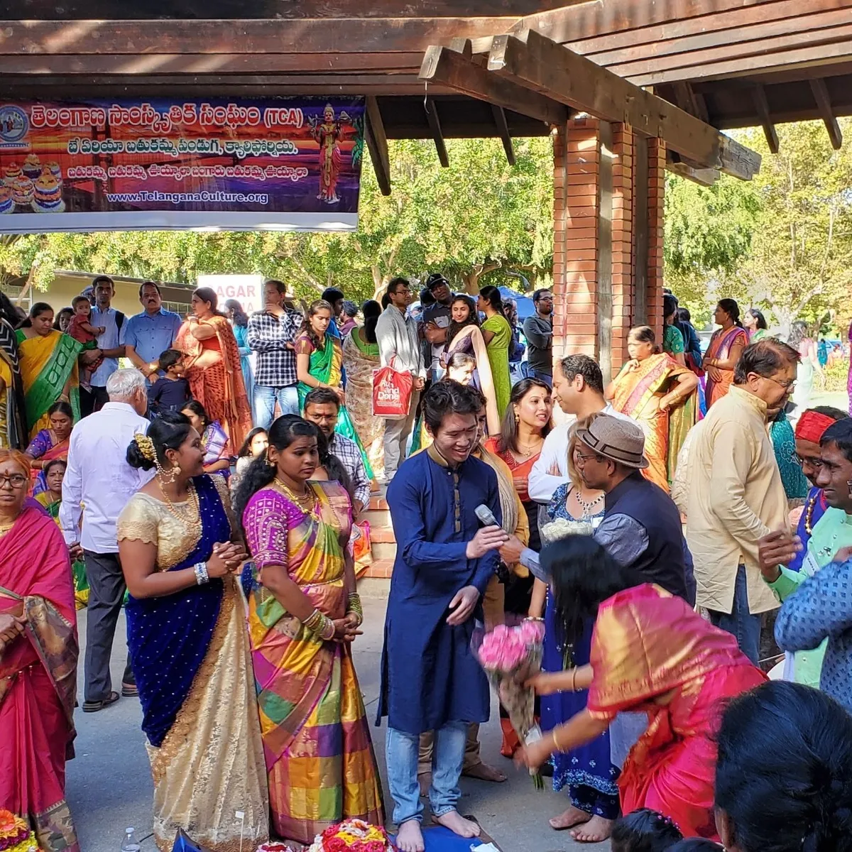 A crowd in saris and kurtas under a shelter, flowers being arranged on the ground, beneath a Telangana Cultural Association banner.